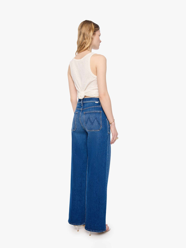 Back view of a woman in a mid blue super high-rise jeans with a loose fit, wide leg, patch pockets, zip fly and 30 3/4 inch inseam with a clean hem.