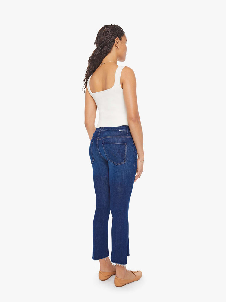 Back view of a petite woman in a dark blue wash high-waisted bootcut is cropped at the ankle with a frayed step-hem jean.