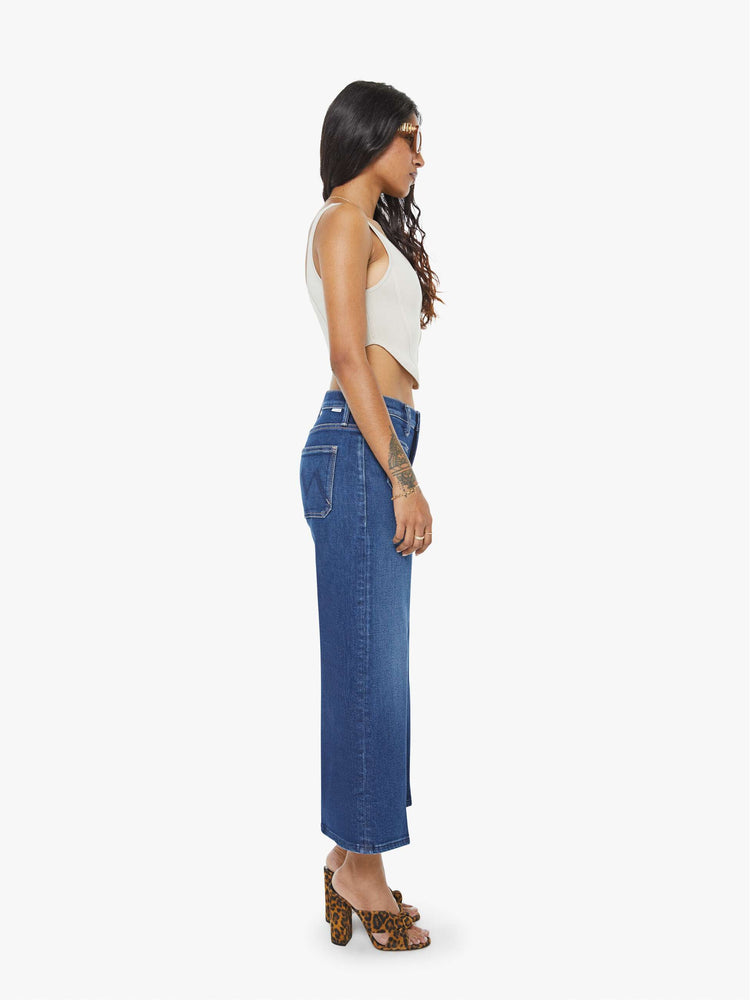 Side view of a woman in a dark blue mid-rise jeans with a loose fit, zip fly, wide leg, patch pockets and ankle-length inseam with a clean hem.
