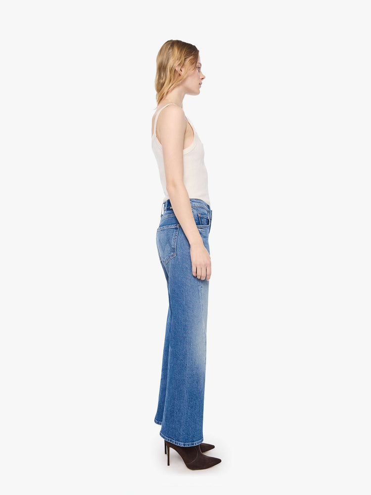 Side view of a petite woman in a mid-blue  mid-rise bootcut with a zip fly, looser leg, twisted outer seam and a long 28-inch inseam with a clean hem. 