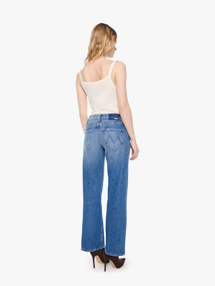 Back view of a petite woman in a mid-blue  mid-rise bootcut with a zip fly, looser leg, twisted outer seam and a long 28-inch inseam with a clean hem. 