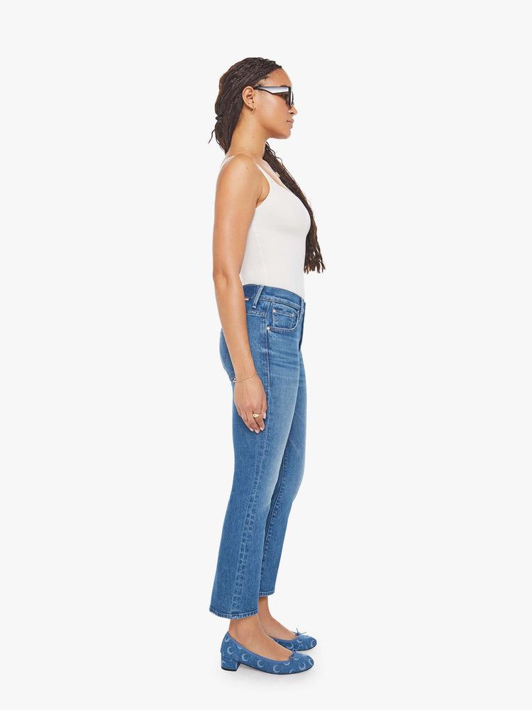 Side view of woman in a mid-blue jean with a high-rise bootcut has an ankle-length inseam, zip fly and a clean hem.