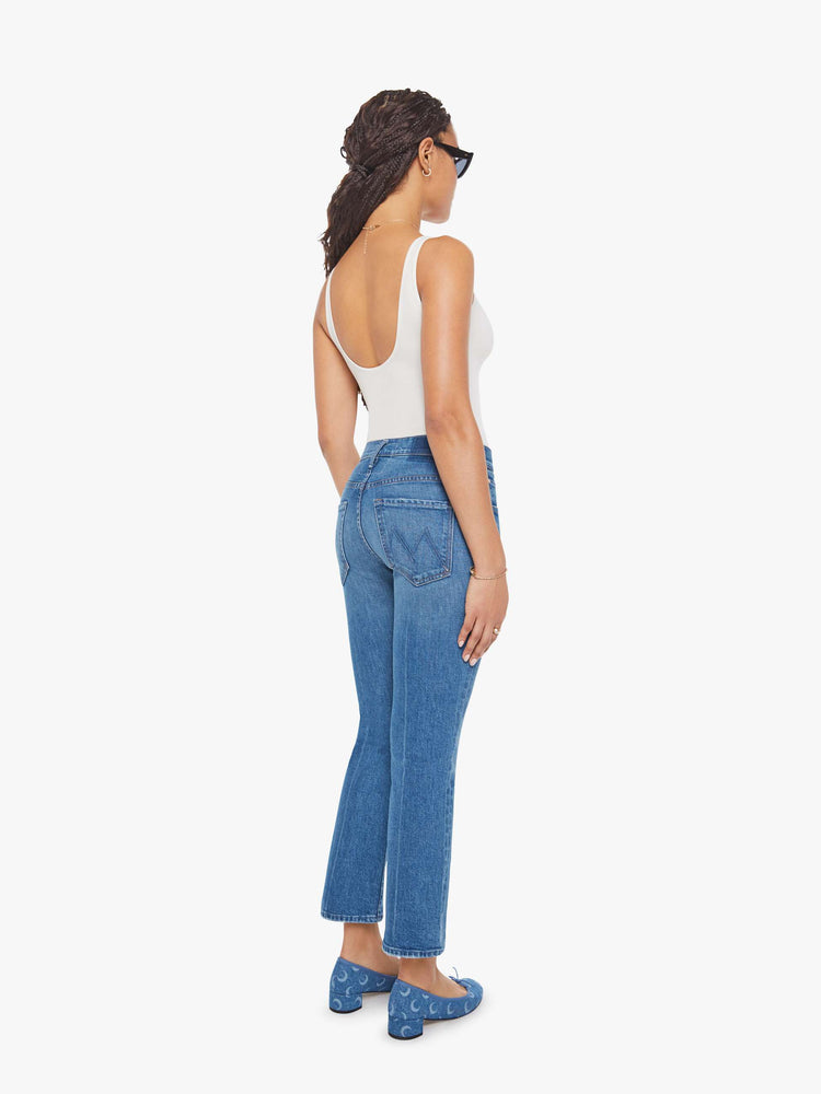 Back view of woman in a mid-blue jean with a high-rise bootcut has an ankle-length inseam, zip fly and a clean hem.