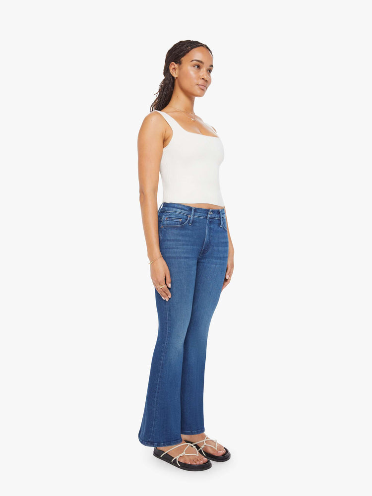 Side angle view of a petite woman in a med blue flare jean has a mid rise, zip fly, sneaker-length inseam and a clean hem.