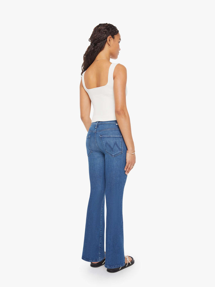 Back view of a petite woman in a med blue flare jean has a mid rise, zip fly, sneaker-length inseam and a clean hem.