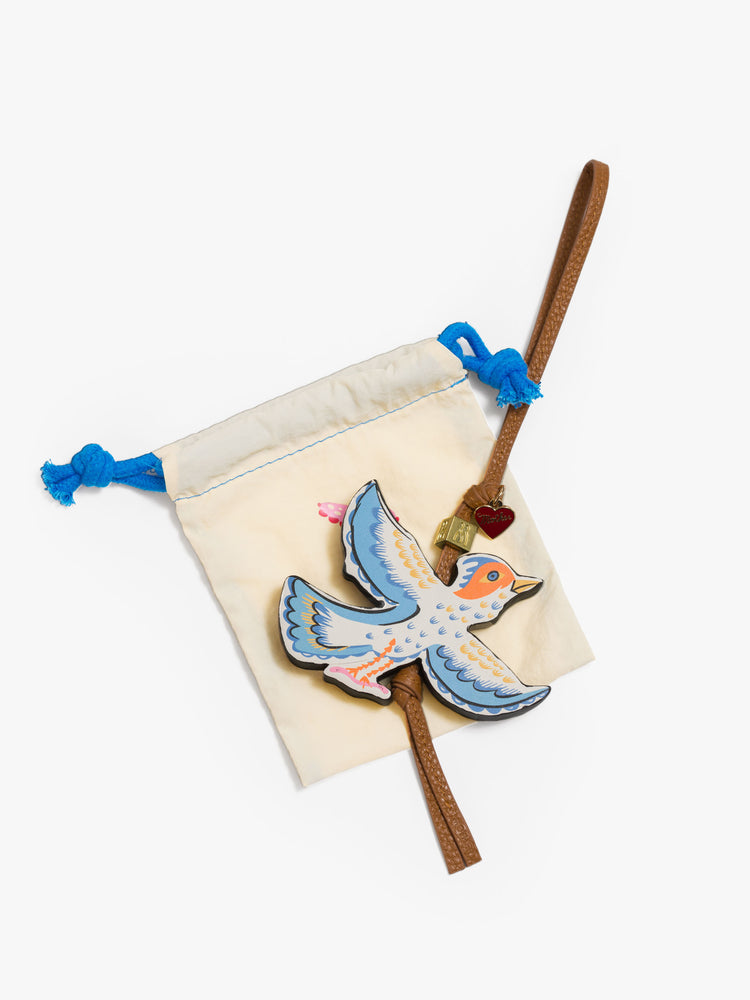 A tan bag charm made from 100% leather with a heart charm, M bead and a colorful 3.7 x 2.7-inch bird at the end sitting on a dust bag,