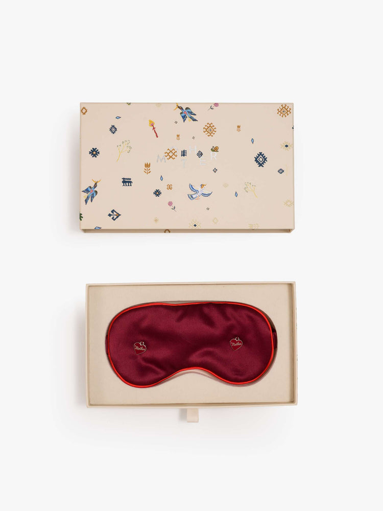 Gift box view of a red sleeping mask with two heart charms on the front.