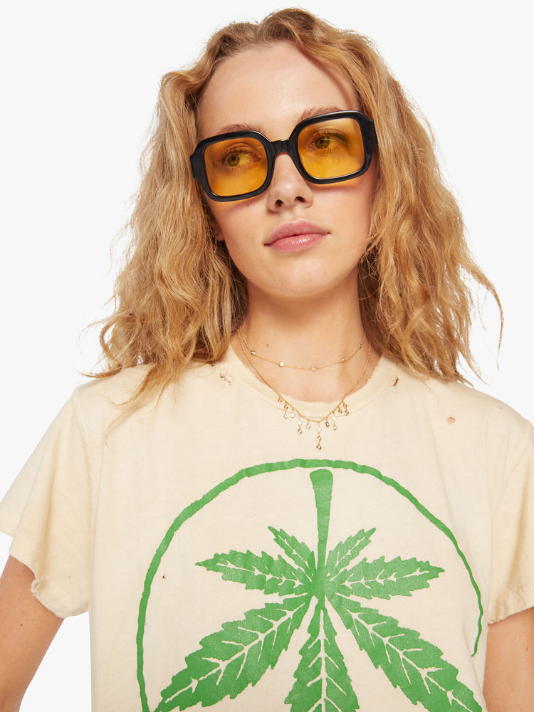 WOMEN Close up view of a woman tea-stained white hue with small tears throughout, the tee offers a sign of peace with a green weed leaf graphic on the front.