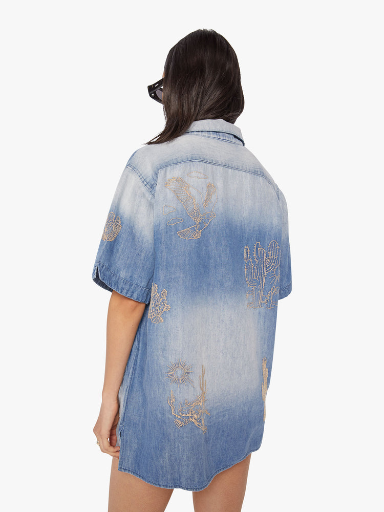 Side back view of a women's short-sleeve collared shirt in a light blue hue with contrast fading and embroidered desert creatures throughout.