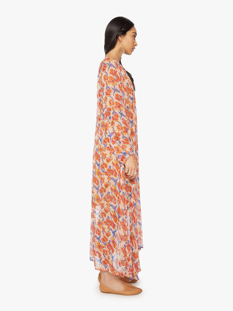Side full body view of a woman's maxi dress in off-white chiffon with a watercolor floral print in orange and navy with voluminous sleeves and has an A-line cut.