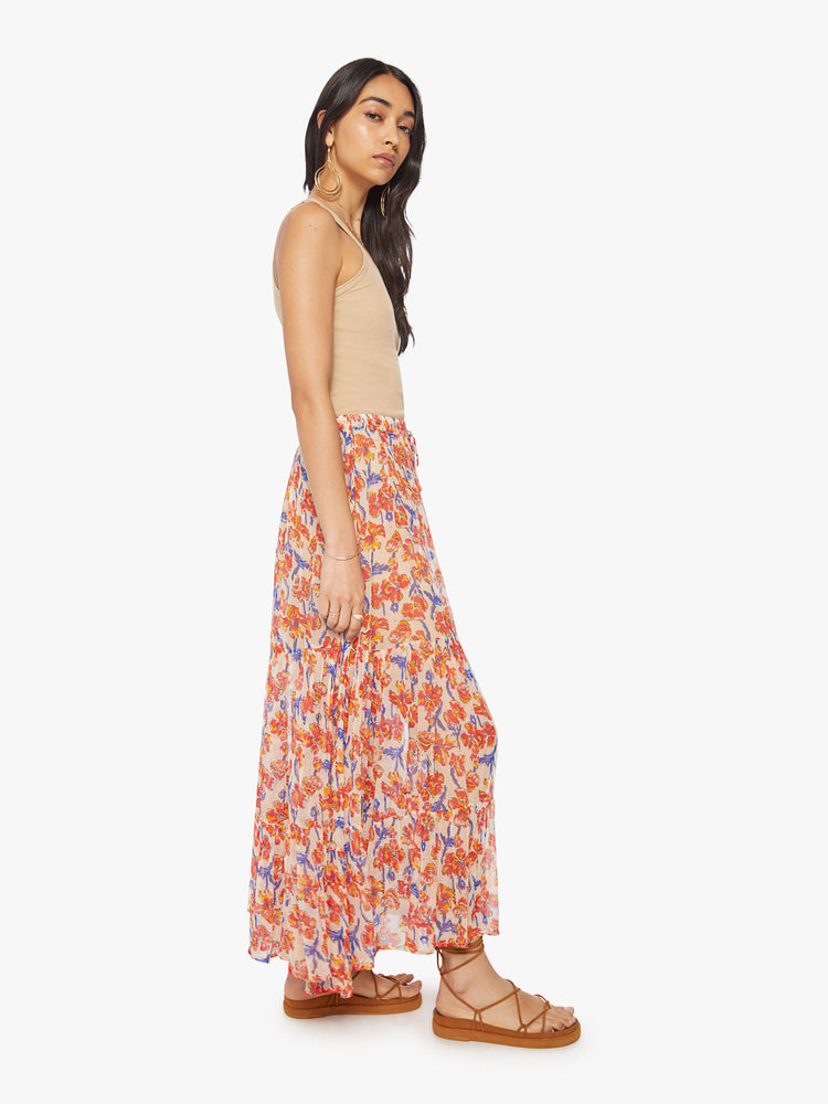 Side view of a woman maxi skirt in an off-white chiffon with a orange/navy watercolor floral print with a drawstring waistband and ruffled hem.