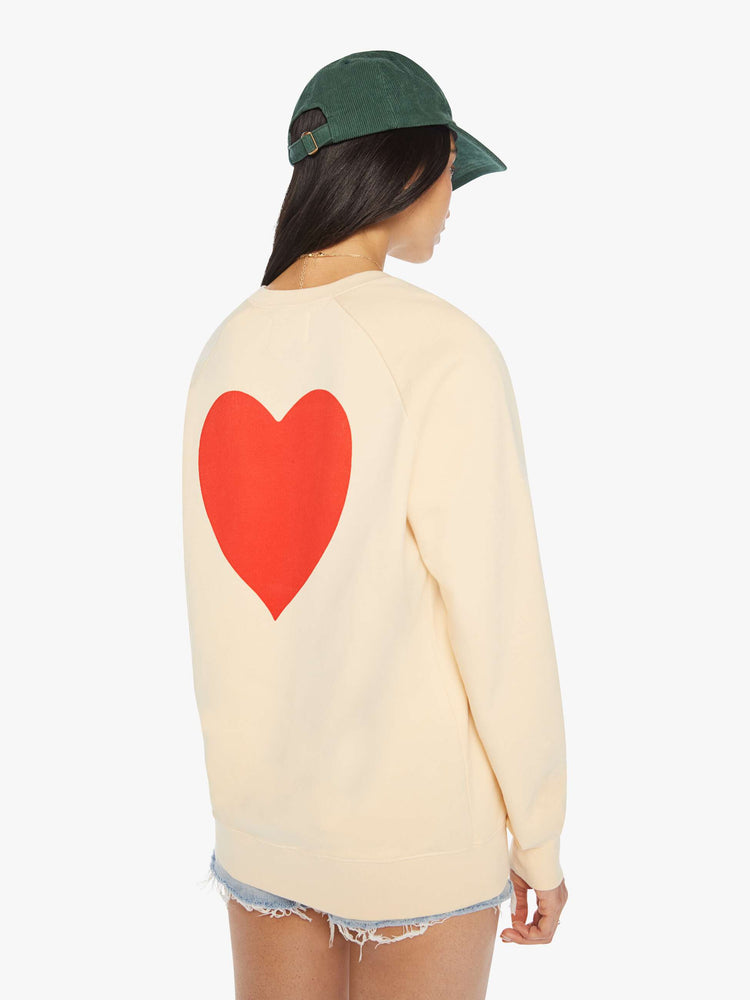 Back view of a woman crewneck sweatshirt in a cream hue with raglan sleeves and is decorated with a red heart graphic on the back and a logo on the front.