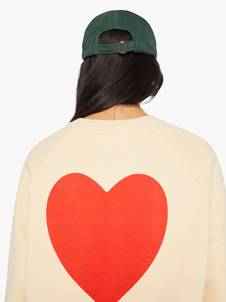 Back close up view of a woman crewneck sweatshirt in a cream hue with raglan sleeves and is decorated with a red heart graphic on the back and a logo on the front.