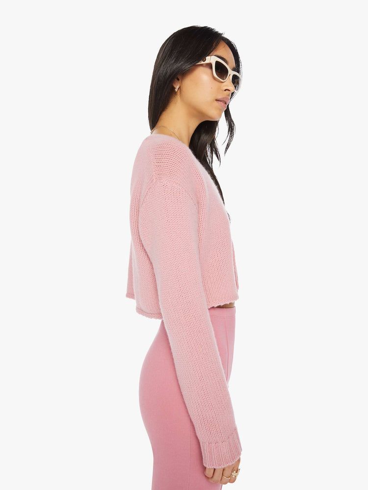 Side view of a woman pink cropped sweater with a V-neck, buttons down the front, drop shoulders and a cropped fit.