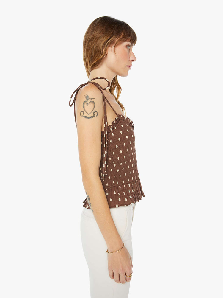 Side view of a woman brown and white polka dots blouse with tied spaghetti straps, a V-neck, smocked bodice and ruffled trim.