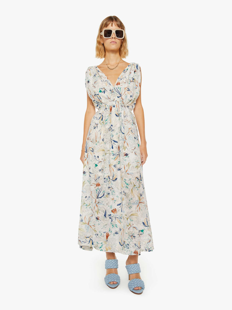 Front view of a woman in a maxi dress with a V-neck, wide straps, a gathered waist and a loose, flowy skirt with an ankle-length hem in off white with floral print.