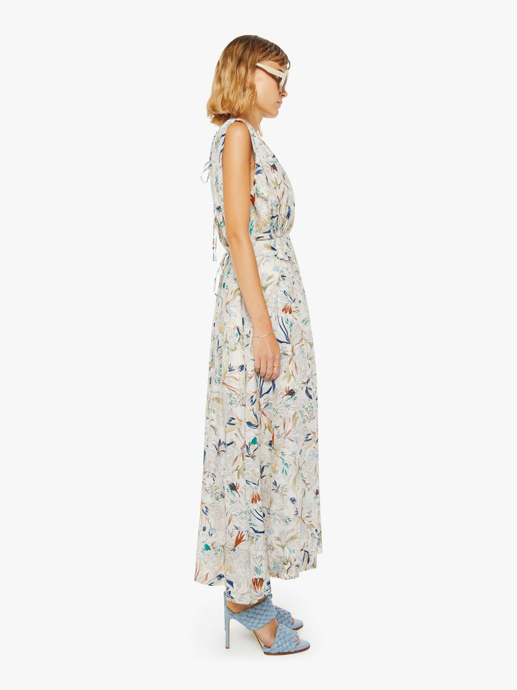 Side view of a woman in a maxi dress with a V-neck, wide straps, a gathered waist and a loose, flowy skirt with an ankle-length hem in off white with floral print.