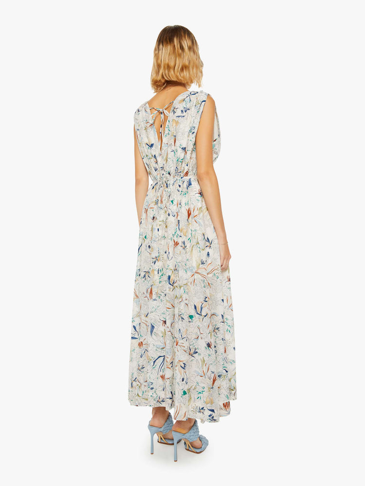Back view of a woman in a maxi dress with a V-neck, wide straps, a gathered waist and a loose, flowy skirt with an ankle-length hem in off white with floral print.