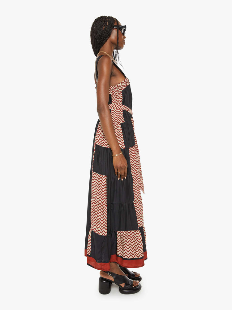 Side view of a woman color blocked in black and a red-and-white zig zag print, and features a square neck, detailed straps, optional waist sash and an ankle-grazing hem.