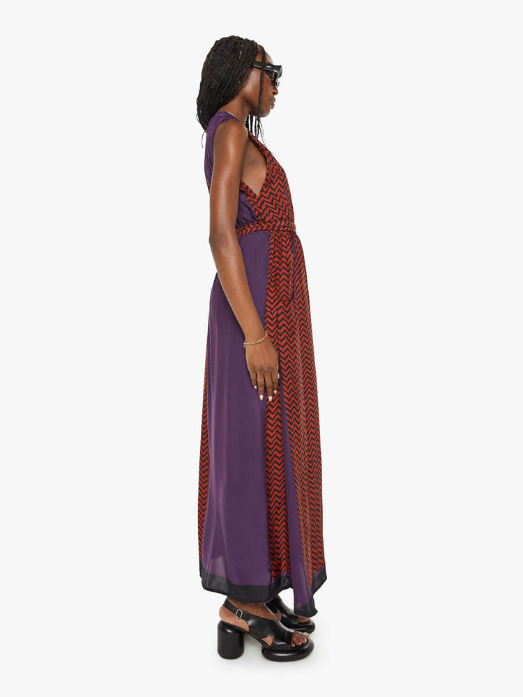Side view of a woman purple and red zig zag print with a maxi dress V-with a neck, fitted waist that ties and ankle-length hem.