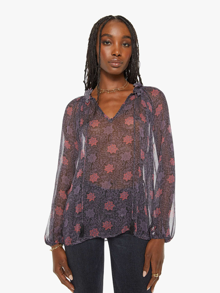 Front view of a woman top in a sheer purple fabric with red floral print and a top has a keyhole neckline with a tasseled tie closure, long balloon sleeves and pleats for a loose fit.