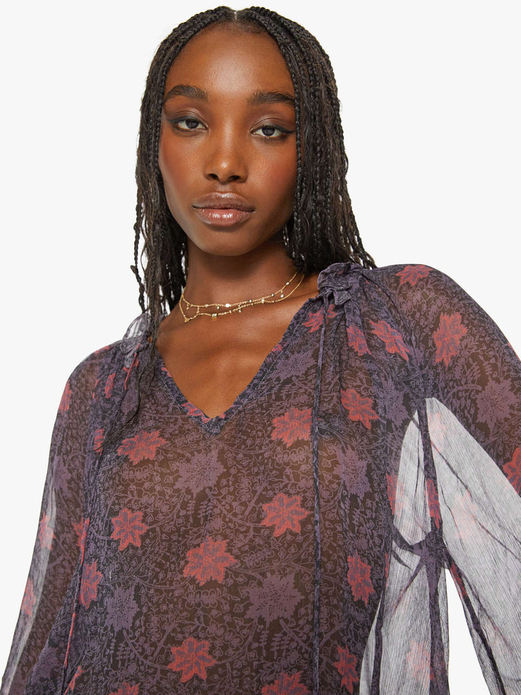 Close up view of a woman top in a sheer purple fabric with red floral print and a top has a keyhole neckline with a tasseled tie closure, long balloon sleeves and pleats for a loose fit.