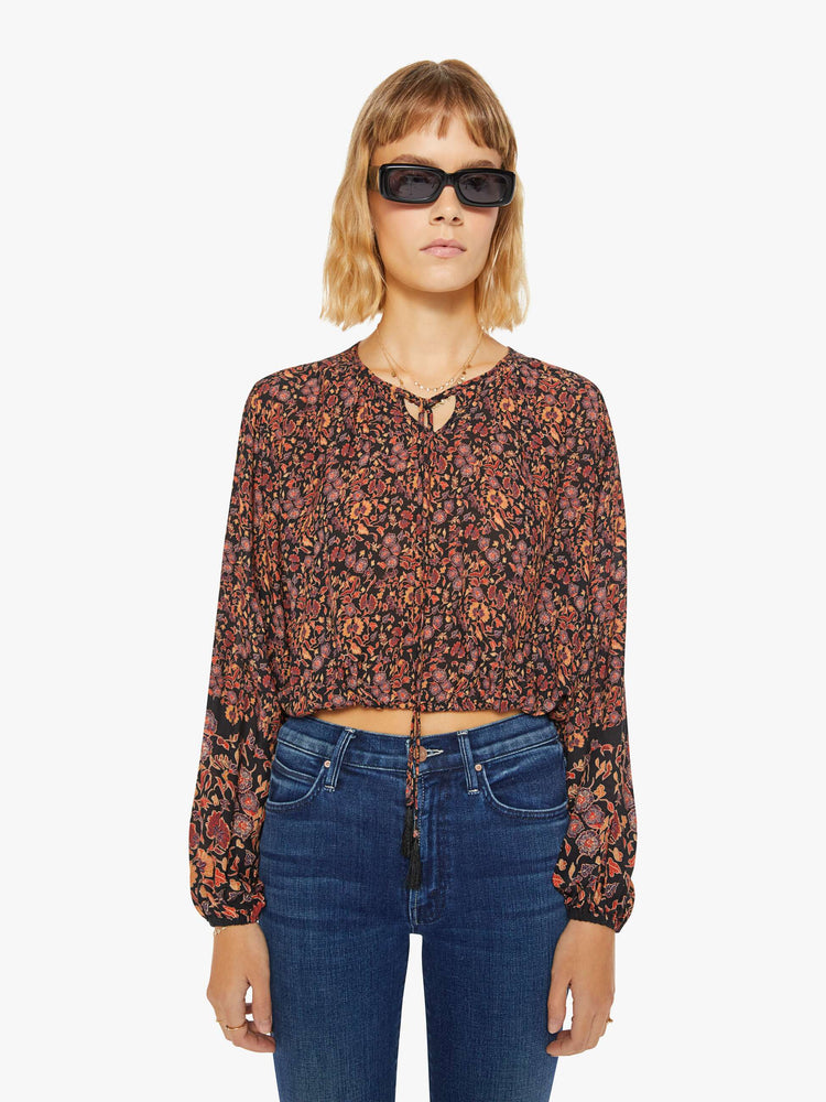 Front view of a woman top in black toned floral print with a keyhole neckline with a tasseled tie closure, drop shoulders, long balloon sleeves and a cropped elastic hem.