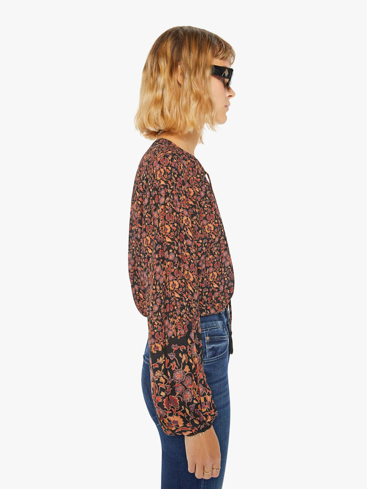 Side view of a woman top in black toned floral print with a keyhole neckline with a tasseled tie closure, drop shoulders, long balloon sleeves and a cropped elastic hem.