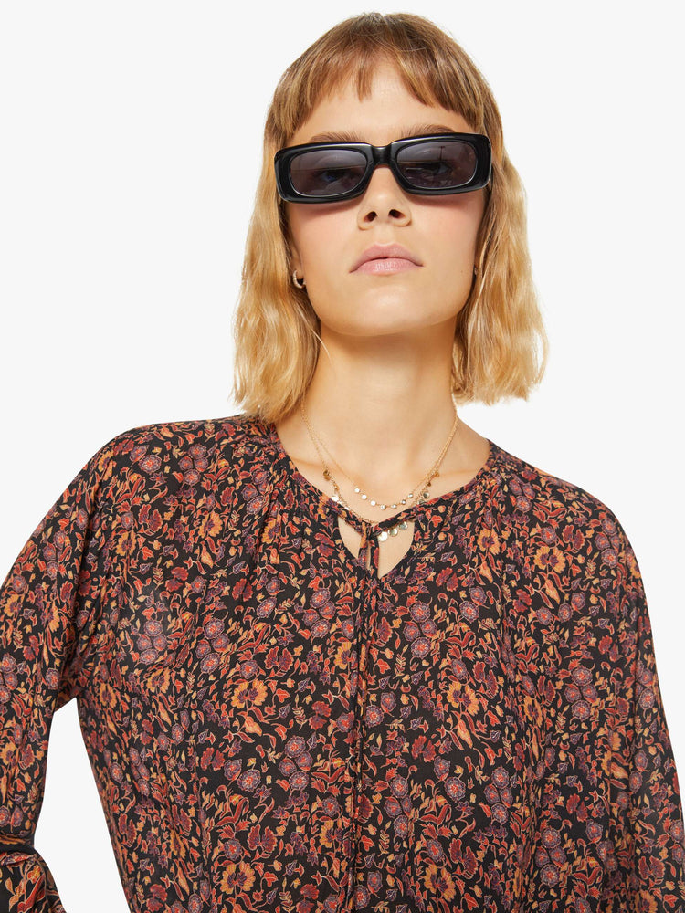 Close up view of a woman top in black toned floral print with a keyhole neckline with a tasseled tie closure, drop shoulders, long balloon sleeves and a cropped elastic hem.