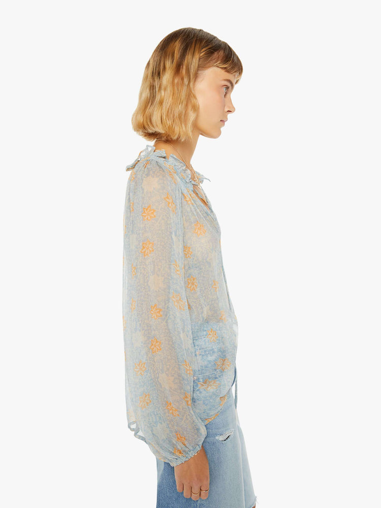 Side view of a woman in a sheer baby-blue fabric with a tangerine floral print shirt with a keyhole neckline with a tasseled tie closure, long balloon sleeves and a loose fit.