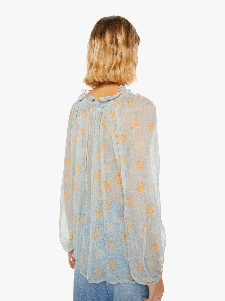 Back view of a woman in a sheer baby-blue fabric with a tangerine floral print shirt with a keyhole neckline with a tasseled tie closure, long balloon sleeves and a loose fit.