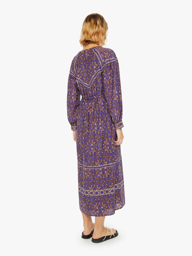 Back view of a woman maxi dress in a maroon, purple and cream print with a V-neck, long balloon sleeves, a tied waist and flowy skirt.