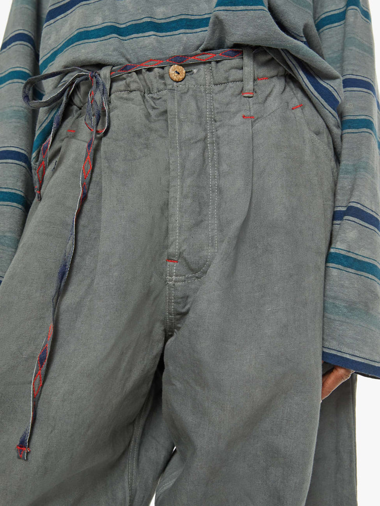 Close up view of a woman army-green hue with a mid rise with pleats at the waist for an even wider fit, an extra-long inseam that puddles at the hem and a colorful woven belt that ties.