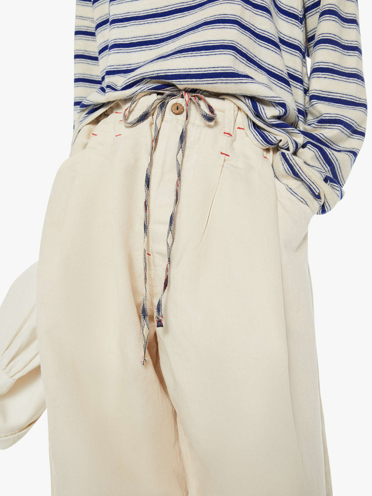 Close up view of a woman creamy hue wide leg with a mid rise with pleats at the waist for an even wider fit, an extra-long inseam that puddles at the hem and a colorful woven belt that ties.