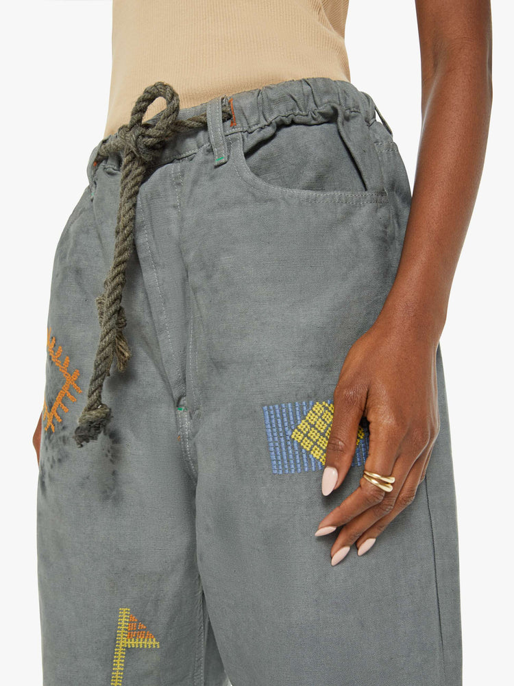 Close up view of a woman army green with color embroidery with a view of a woman in a mid blue wash with colorful embroidery with a mid rise, straight leg, drawstring waist with a woven belt and a loose fit.