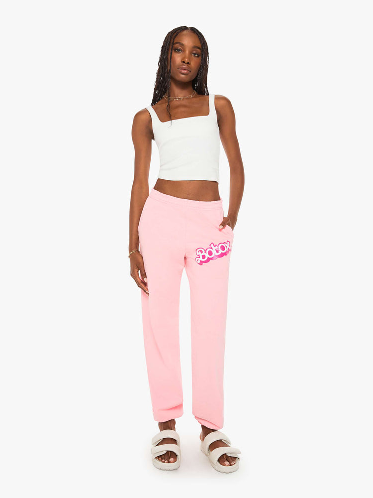 Front view of a woman sweatpants in baby pink have an elastic waist and cuffs, a slightly oversized fit and a two-toned text graphic on the thigh and back.