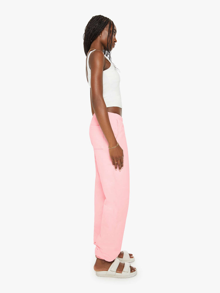 Side view of a woman sweatpants in baby pink have an elastic waist and cuffs, a slightly oversized fit and a two-toned text graphic on the thigh and back.