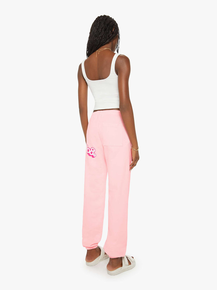 Back view of a woman sweatpants in baby pink have an elastic waist and cuffs, a slightly oversized fit and a two-toned text graphic on the thigh and back.