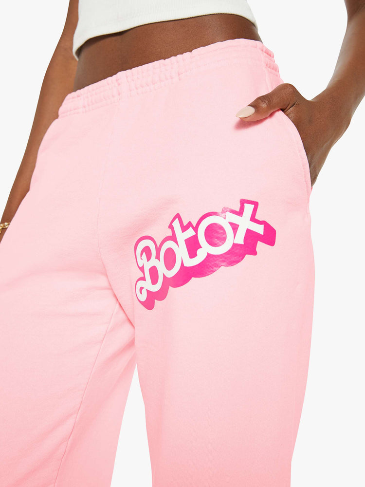 Waist close up view of a woman sweatpants in baby pink have an elastic waist and cuffs, a slightly oversized fit and a two-toned text graphic on the thigh and back.