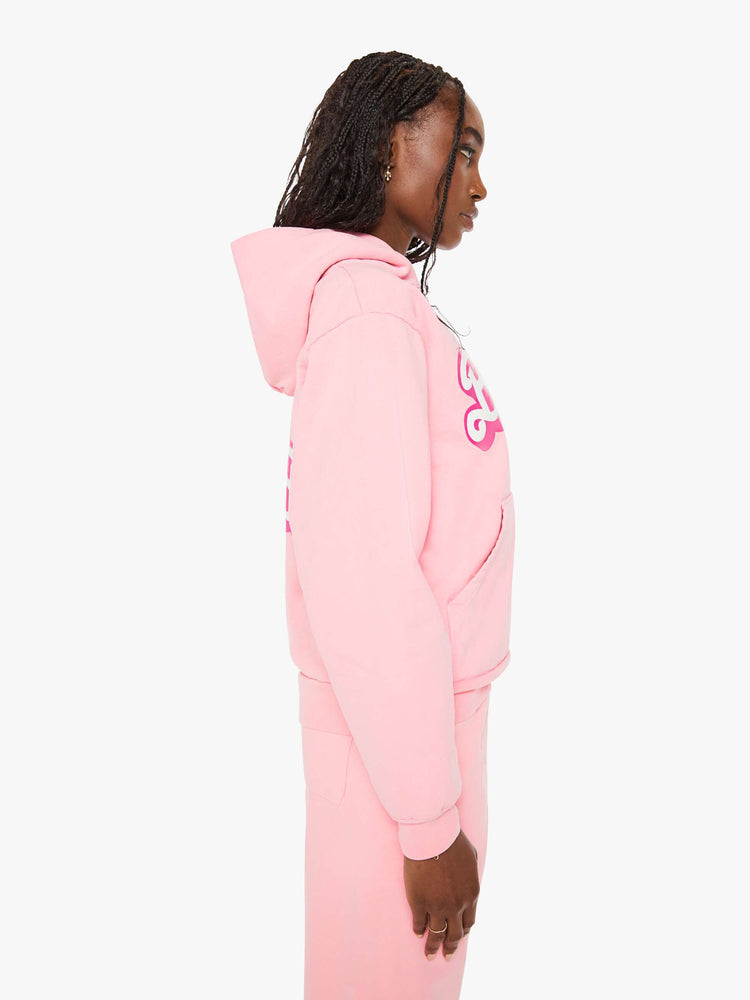 Side view of a woman sweatshirt in baby pink has a front patch pocket, ribbed hems and a slightly oversized fit with a two-toned text graphic on the front.