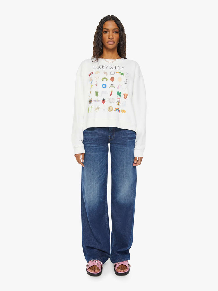 Full body view of a woman white long sleeve sweatshirt features a colorful hand-drawn graphic with a variety of good luck charms.