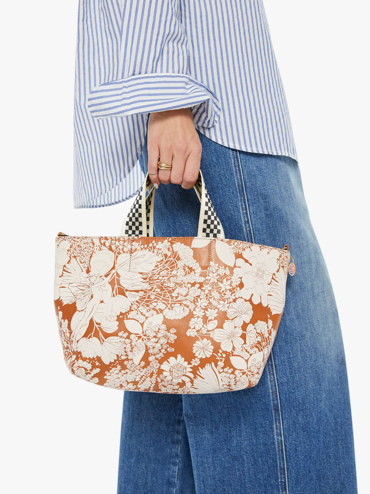 In model hand view of a rectangular bag features a zip closure and checkered top handle, and is designed in French Fairy Tale, a brown and white floral print. Pair it with the Link Up strap for added versatility.