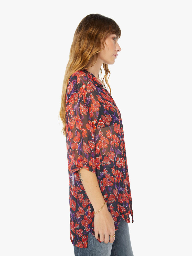 Side view of a woman in a black and red floral print, and features a V-neck, drop shoulders, elbow-length sleeves, buttons down the front and a long, thigh-grazing hem shirt.