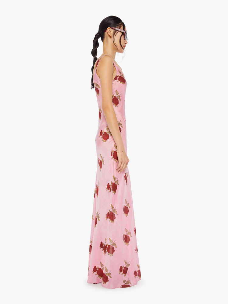 Side view of a woman wearing a pink, sleeveless, maxi dress with red rose print