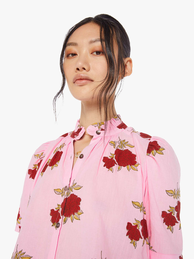 Detail view of a woman wearing a pink button up shirt with red rose print, ruffled collar, and short sleeves