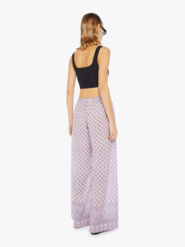 Back view of a woman wearing loose cotton pants with all-over purple floral print and drawstring waist