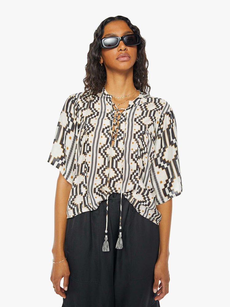 A front view of a woman wearing a black and white pattern short sleeve blouse featuring a laced v-neck with tassels and a flowy fit, paired with a black cargo pant.