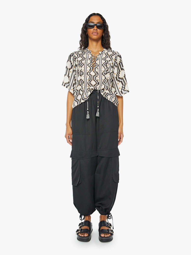 A front full body view of a woman wearing a black and white pattern short sleeve blouse featuring a laced v-neck with tassels and a flowy fit, paired with a black cargo pant.