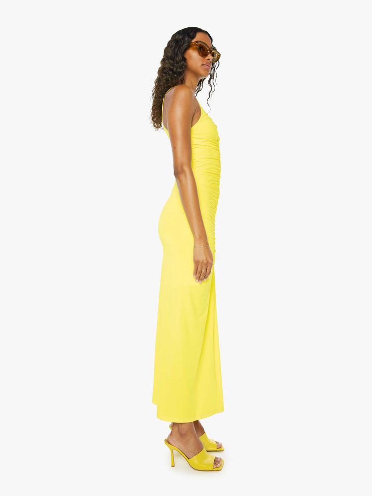 Side view of a woman wearing a bright yellow sleeveless dress with a small v-neck, a long gathered seam down the front, and an ankle length hem.