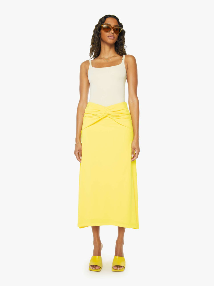 A front view of a woman wearing a bright yellow skirt with a flowy fit, featuring a front knotted wrap detail, paired with a white tank top.
